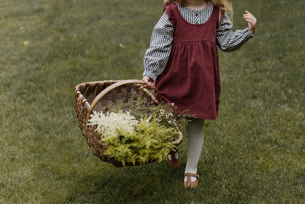 A girl runs through a field holding a basket of dried flowers. She is wearing a Corduroy Pinafore dress and Gingham top.