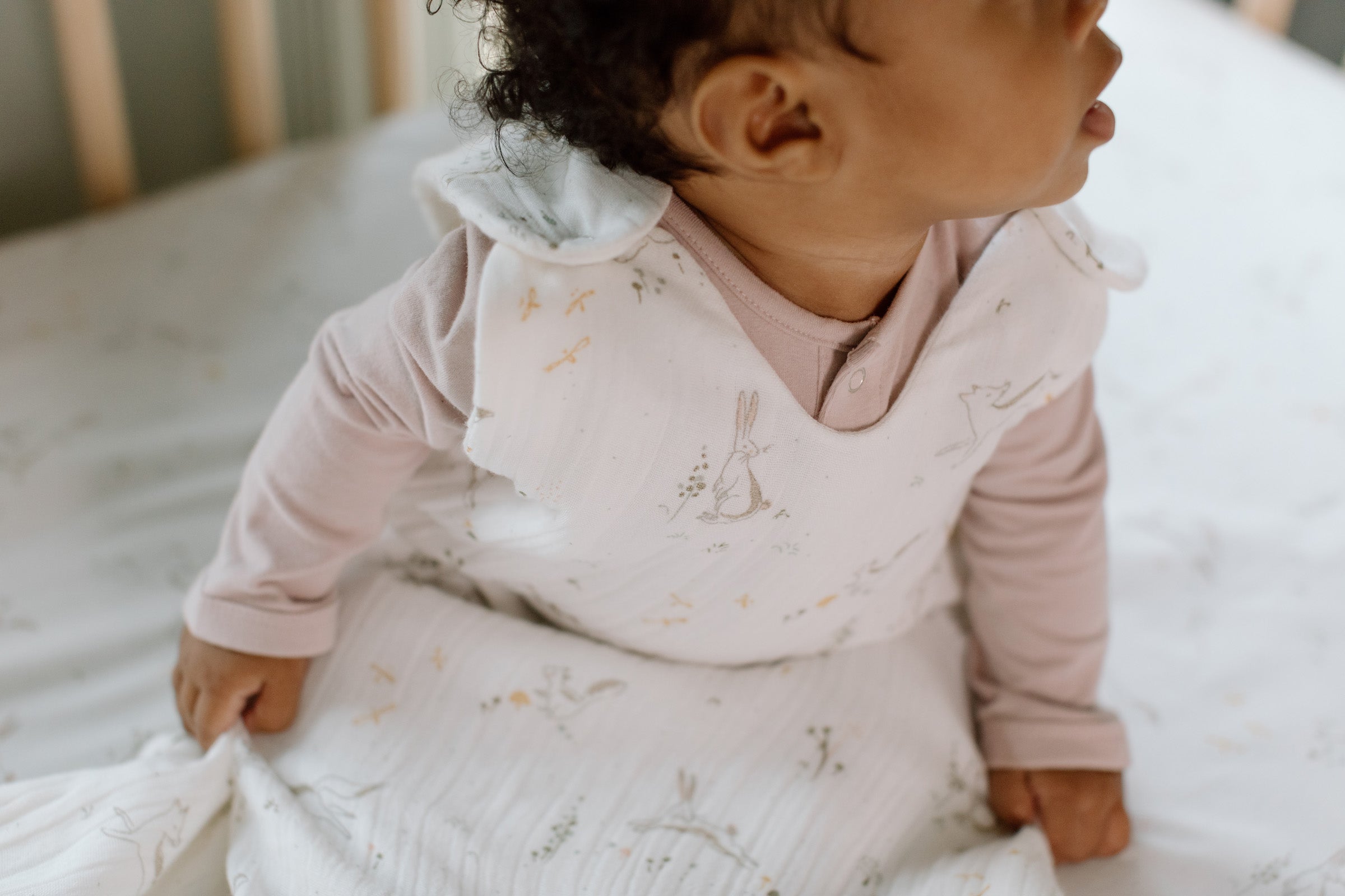 A baby sits in their crib in a sleep bag.