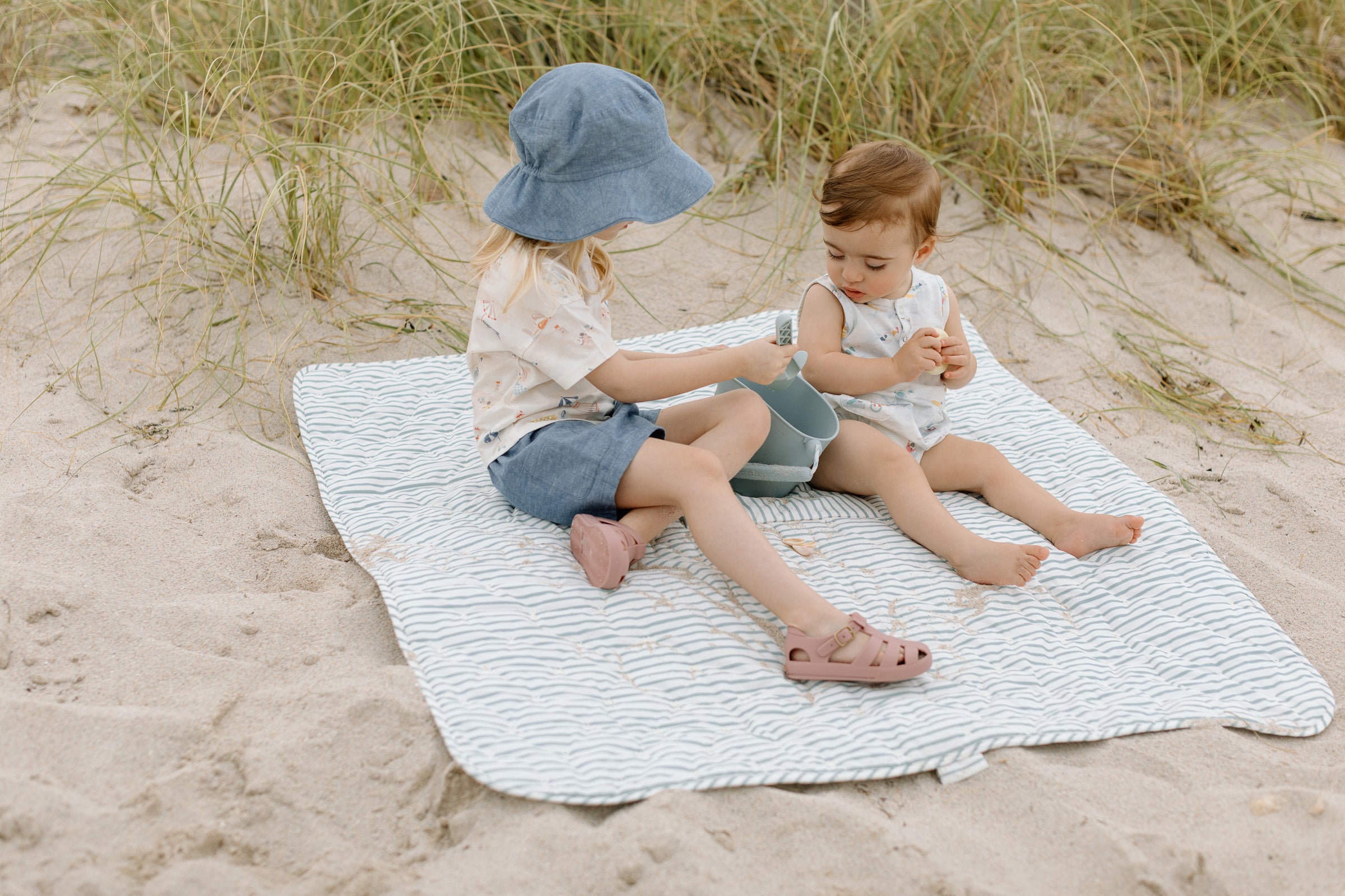 A brother and sister sit on the beach, wearing organic cotton clothing.