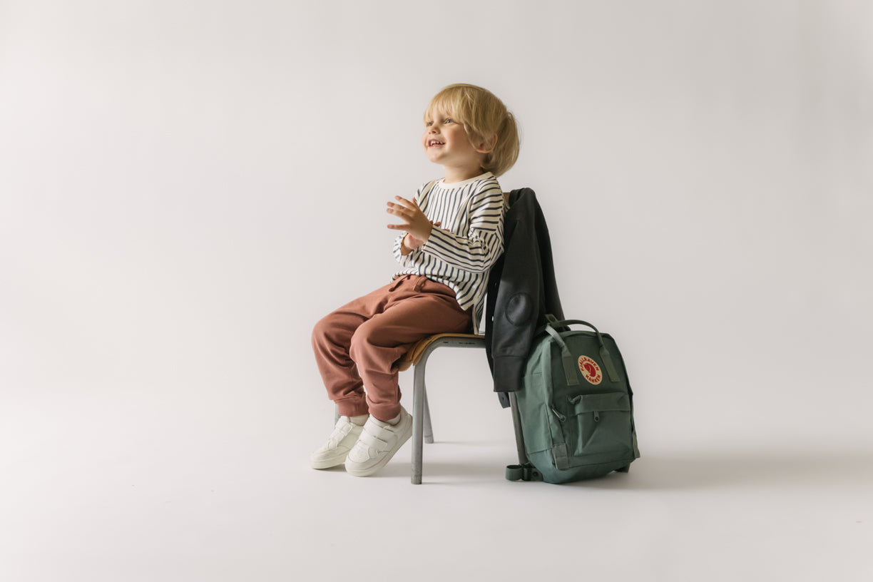 Toddler boy sits on a chair in Pehr French Terry Joggers and long sleeve with a backpack