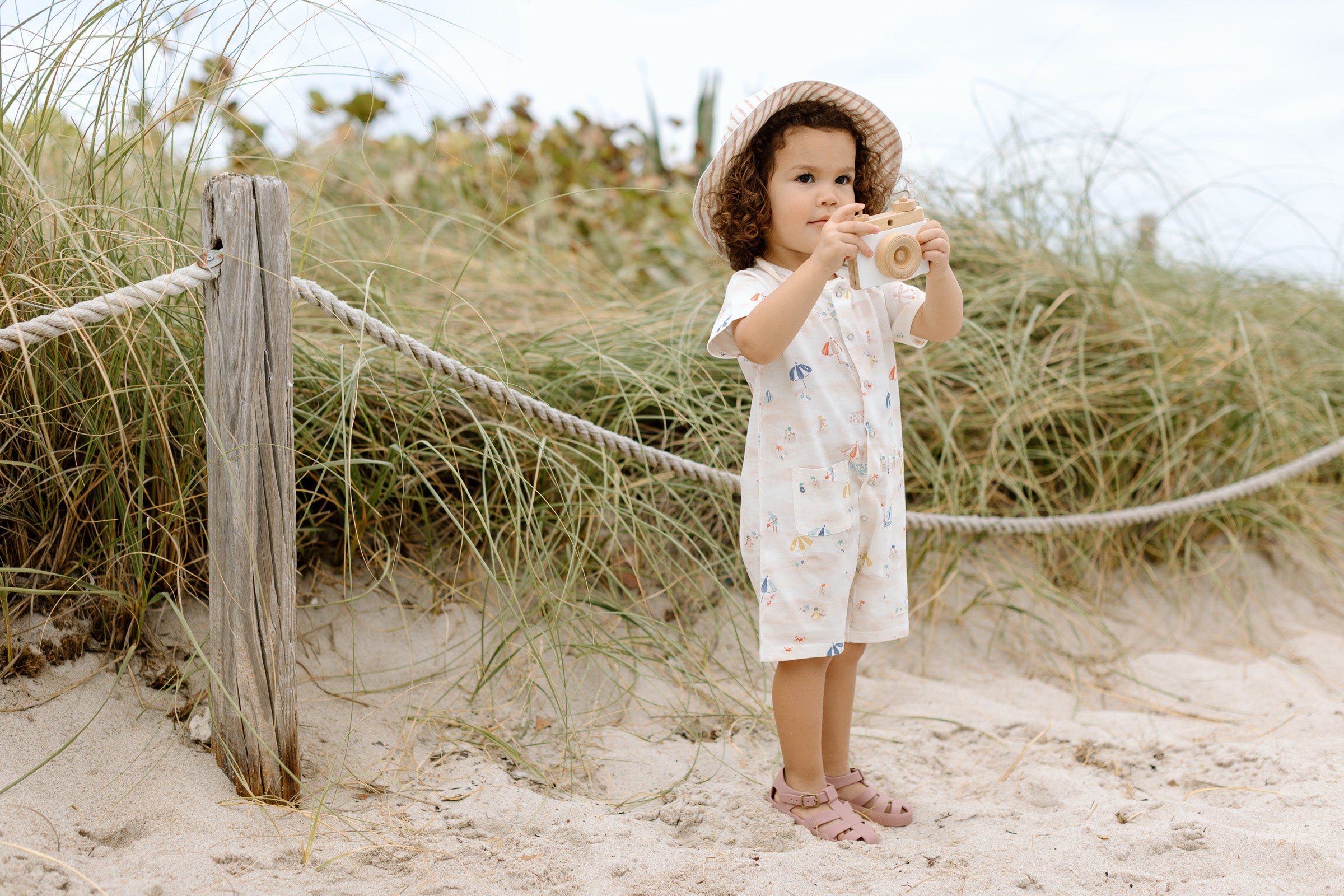 A young girl stands on the beach, holding a wooden camera, wearing a Pehr bucket hat and romper.