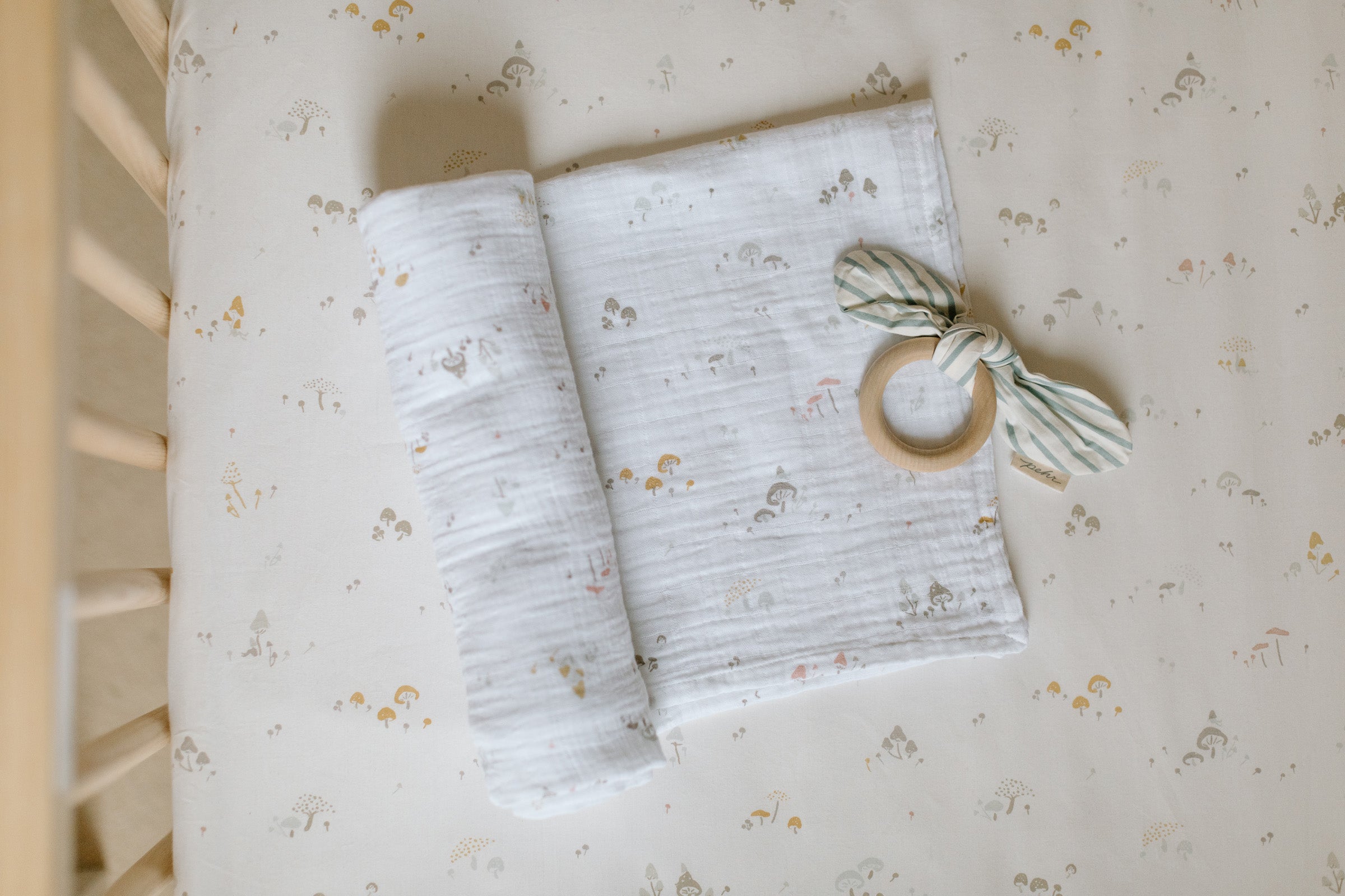 An image of a Pehr crib sheet with a rolled swaddle and On The Go teether.