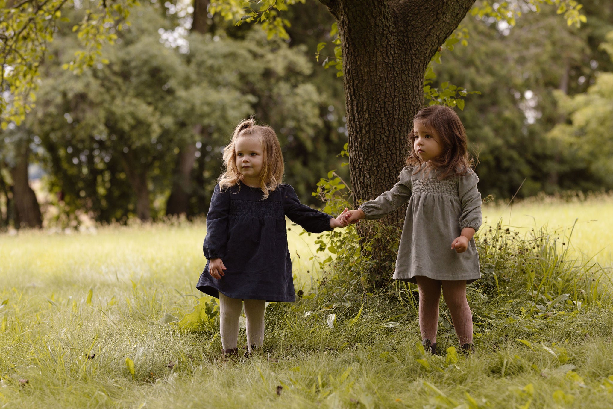 Two girls in corduroy dresses hold hands under a tree. 