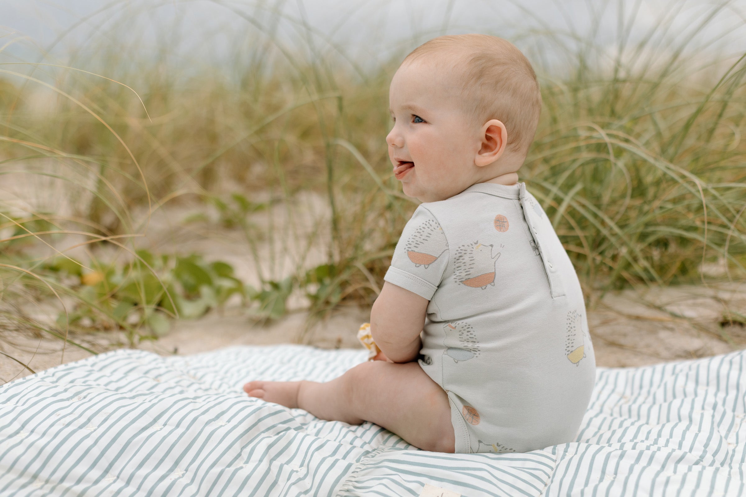 A baby sits on a play mat on the beach, wearing a Park Pets one-piece.