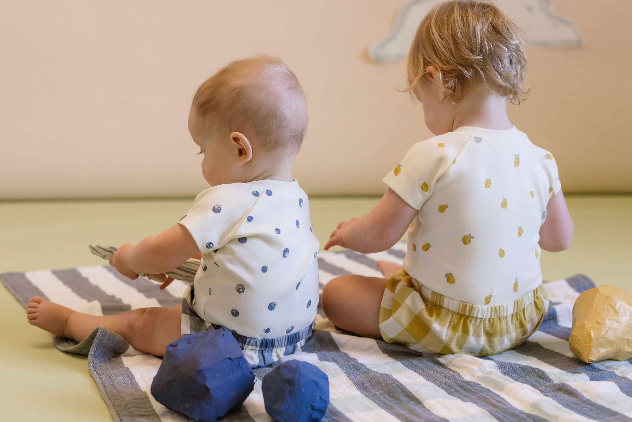 Two children in Fruits one-pieces sit on a blanket. 